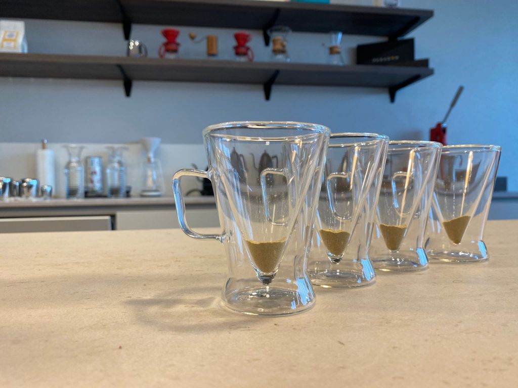 Four double-walled glass mugs on a cafe counter, each with a spoonful of BLISSDBRU powder settled at the base, Chemex brewers softly blurred on the shelf behind