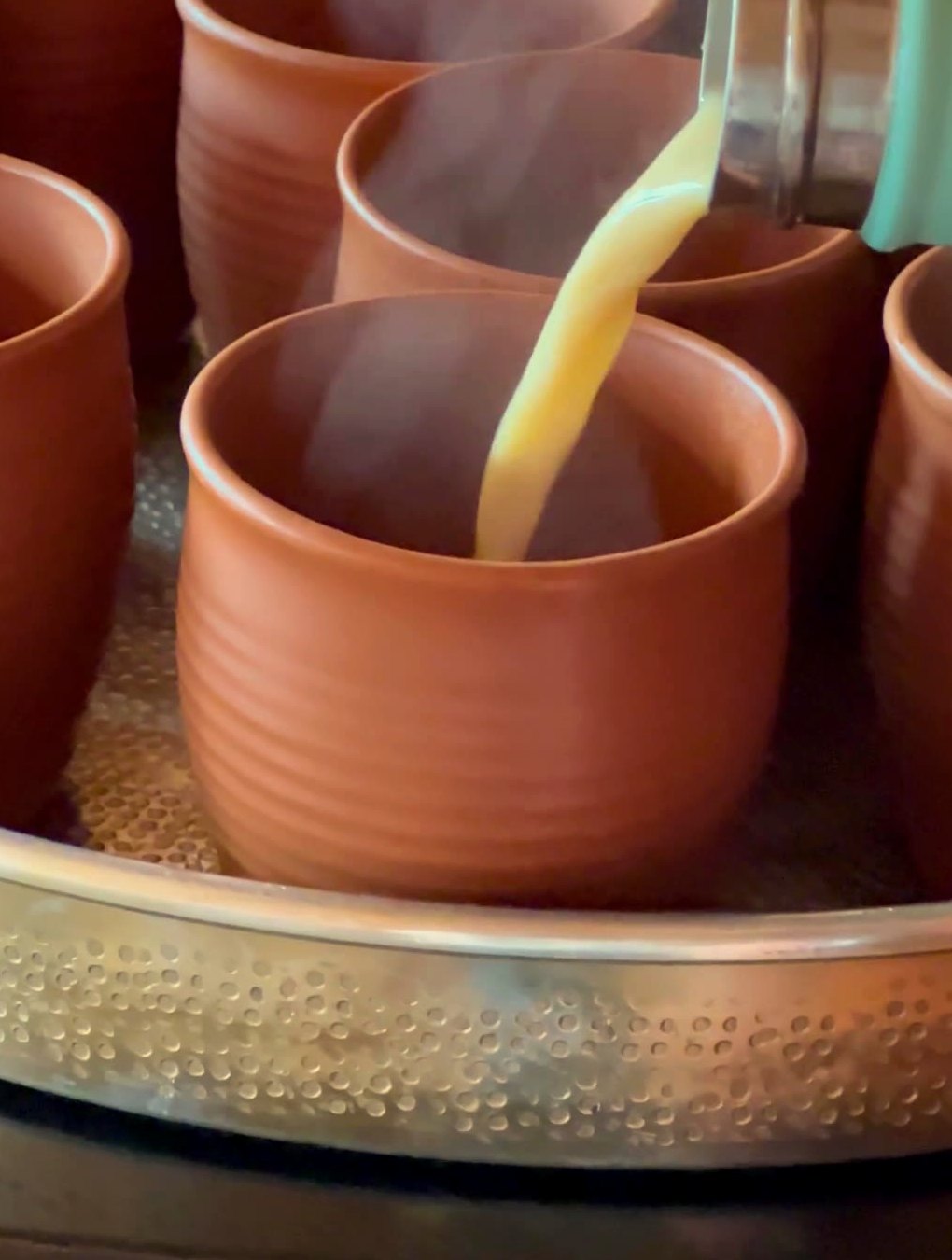 Golden BLISSDBRU being poured, steaming, into terracotta kulhads on a hammered brass tray